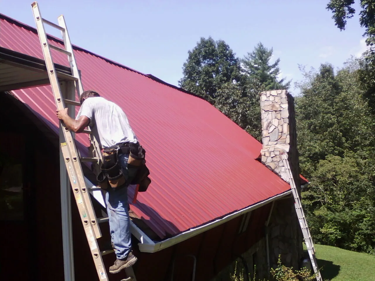 Roofer on a ladder beside a red metal roof and stone chimney — Soffit & Fascia Repair by Roofing Brothers in Donnellson